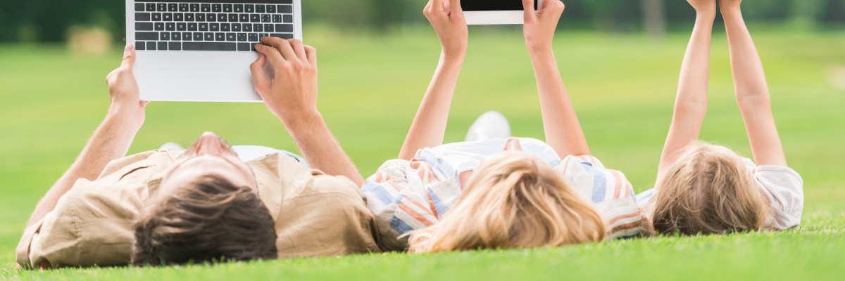 Three people lying on the grass holding up a laptop computer, a tablet and a smart phone