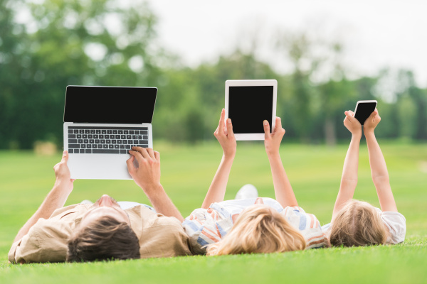 Three people lying on the grass holding up a laptop computer, a tablet and a smart phone