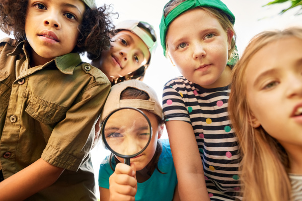 Five curious children with hats on backwards standing and leaning into the frame, with one child looking through a magnifying glass 